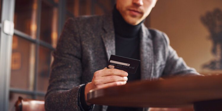 Portrait of modern young guy in formal clothes that sits in the cafe and holds credit card in hand.