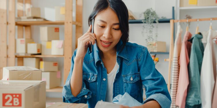 Young Asia businesswoman using smartphone call receiving purchase order and check product on stock work at home office. Small business owner, online market delivery, lifestyle freelance concept.