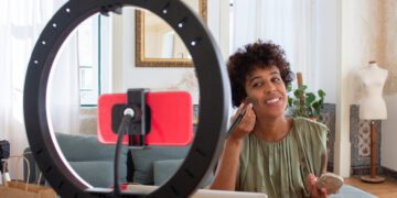 Young Latin woman applying powder on her face for beauty blog. Smiling woman sitting at table in cosy room holding powder box and brush looking at phone camera recording video. Make up and cosmetics blogging concept