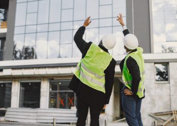 Multi racial builders standing outdoors, back view. Wearing uniform, talking about new glass building. Working on the poject. City infrastructure. Black man.