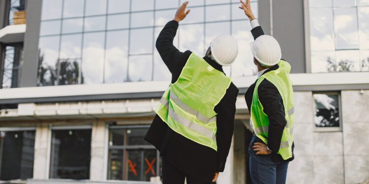 Multi racial builders standing outdoors, back view. Wearing uniform, talking about new glass building. Working on the poject. City infrastructure. Black man.