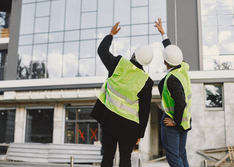 Multi racial builders standing outdoors, back view. Wearing uniform, talking about new glass building. Working on the poject. City infrastructure. Black man.